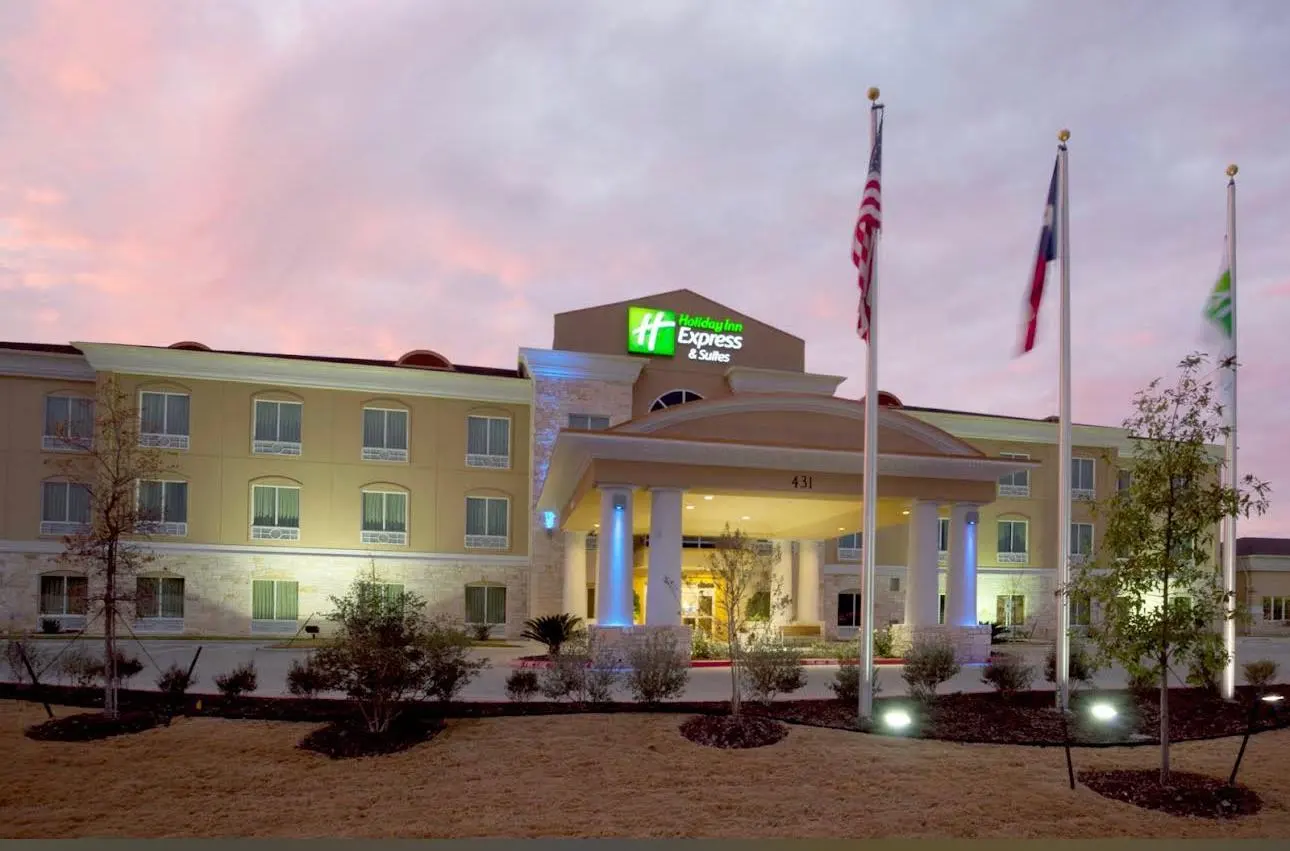 A Holiday Inn Express & Suites at dusk, its columned porte-cochère lit and three flags before it