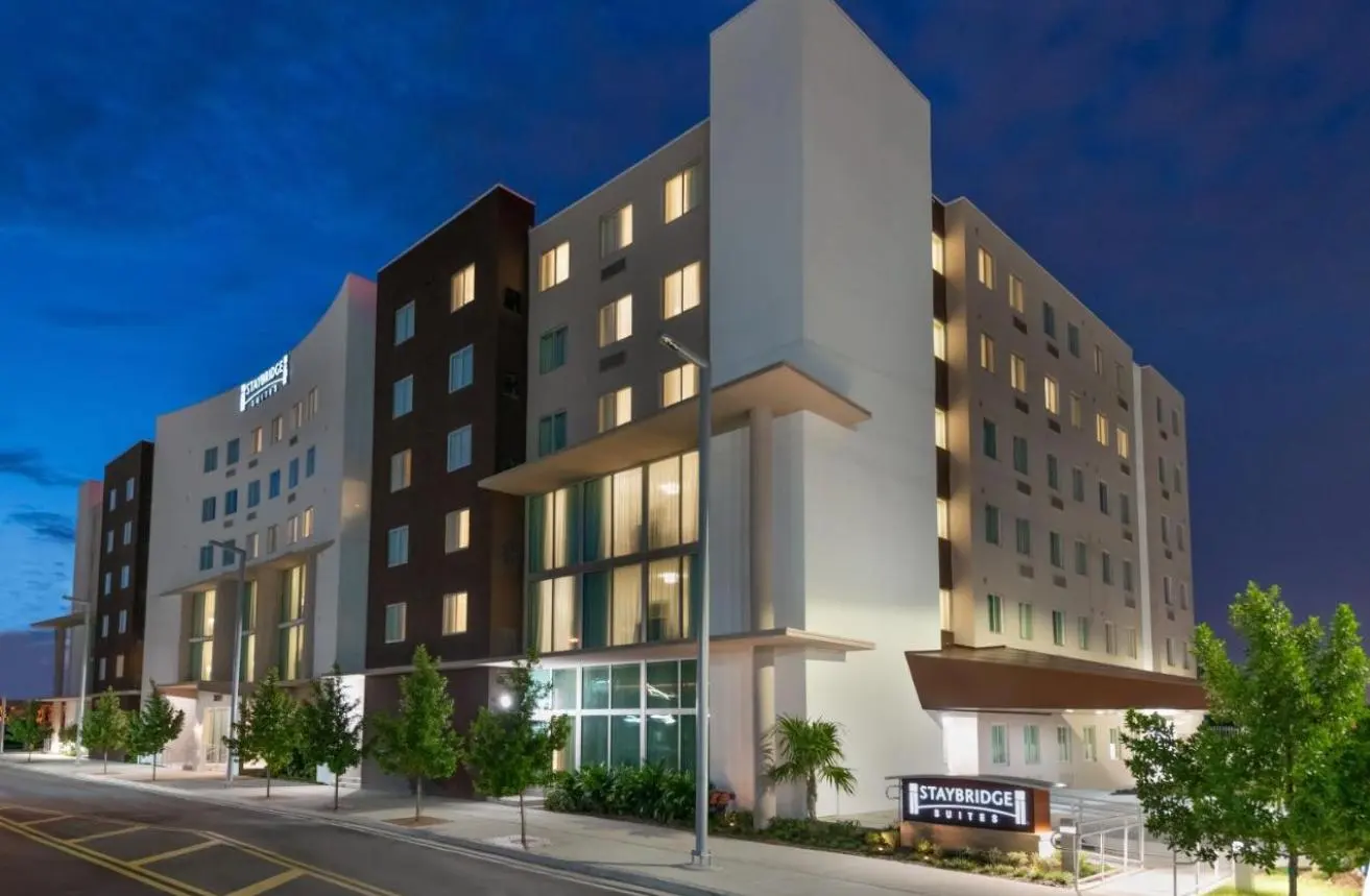 A Staybridge Suites at dusk, its white and dark-brick façade lit from within, the sign at the street